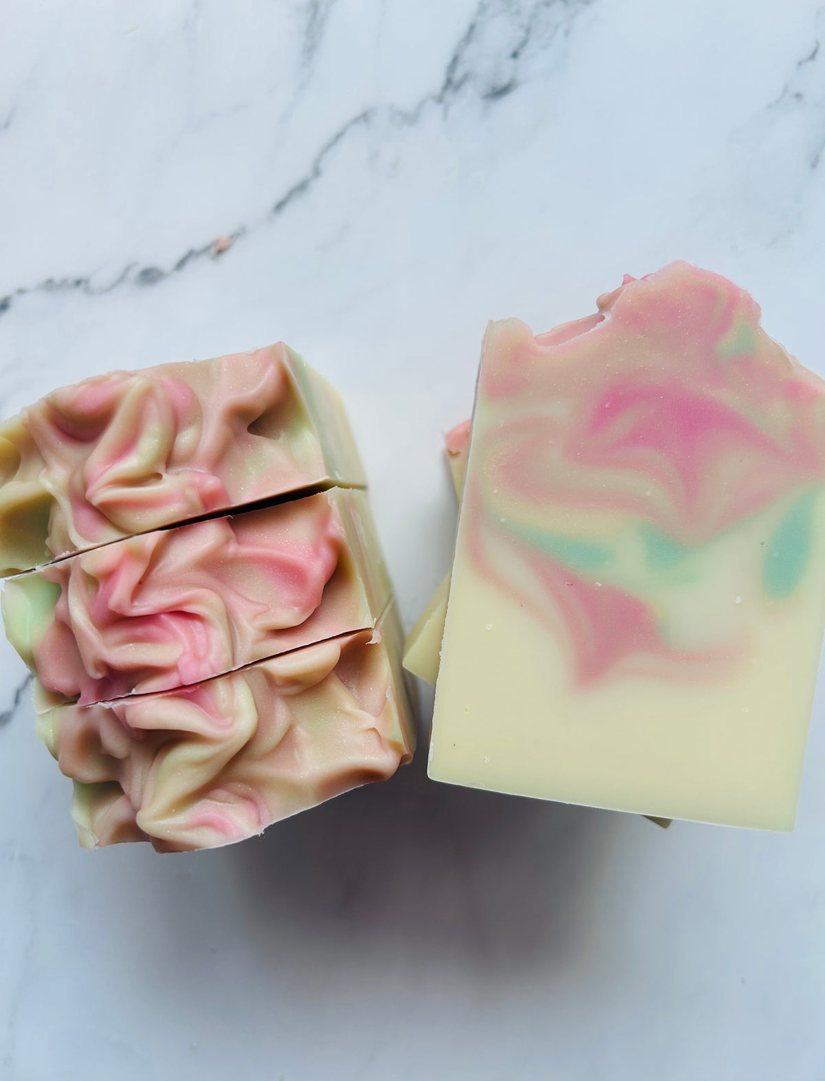 Image of pink, green, and gold swirled soap bars with 3 bars stacked on top of one another and 3 bars standing vertically photographed on a white marble background.