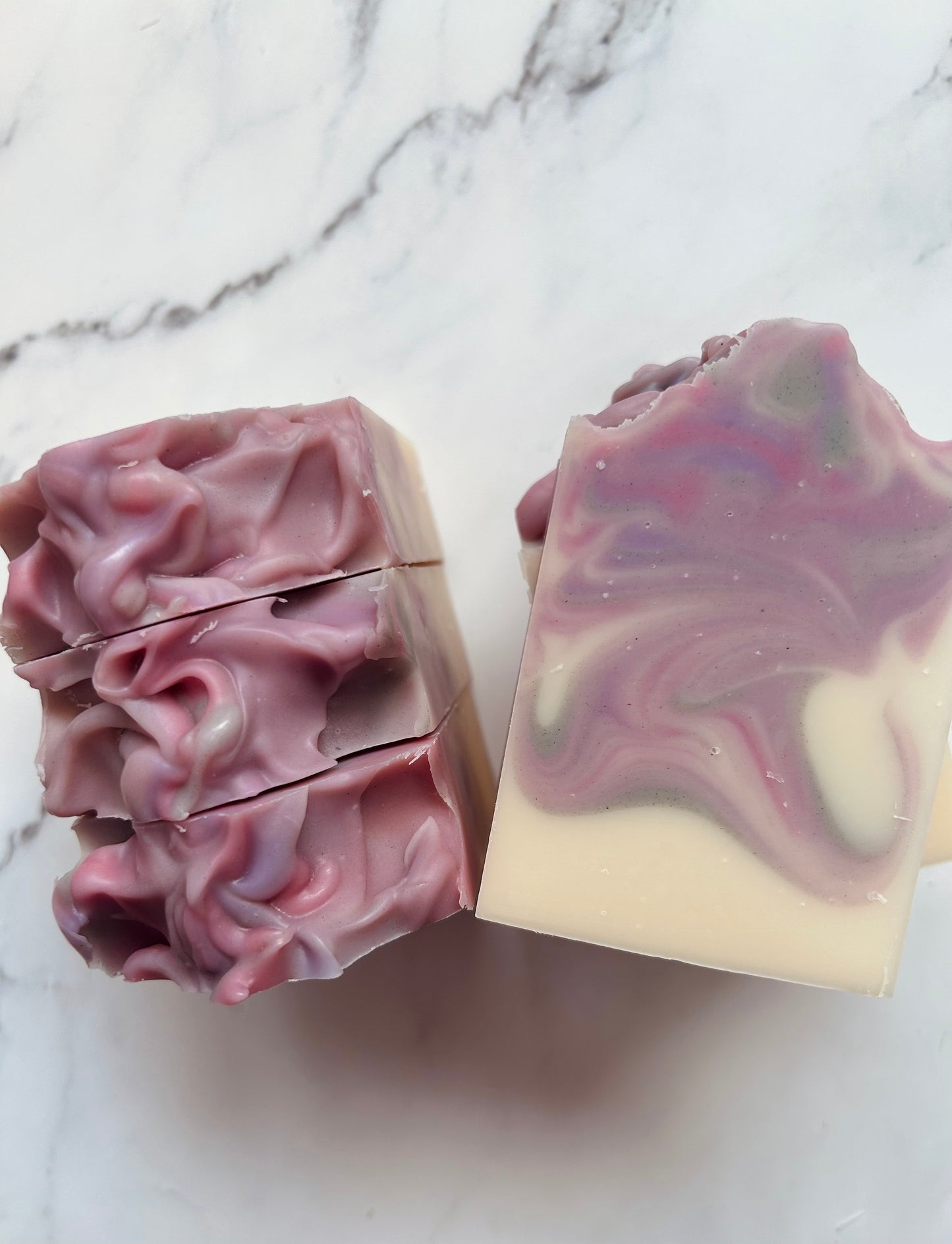 Image of purple, pink, and gray swirled soap bars with 3 bars stacked on top of one another and 3 bars standing vertically photographed on a white marble background.