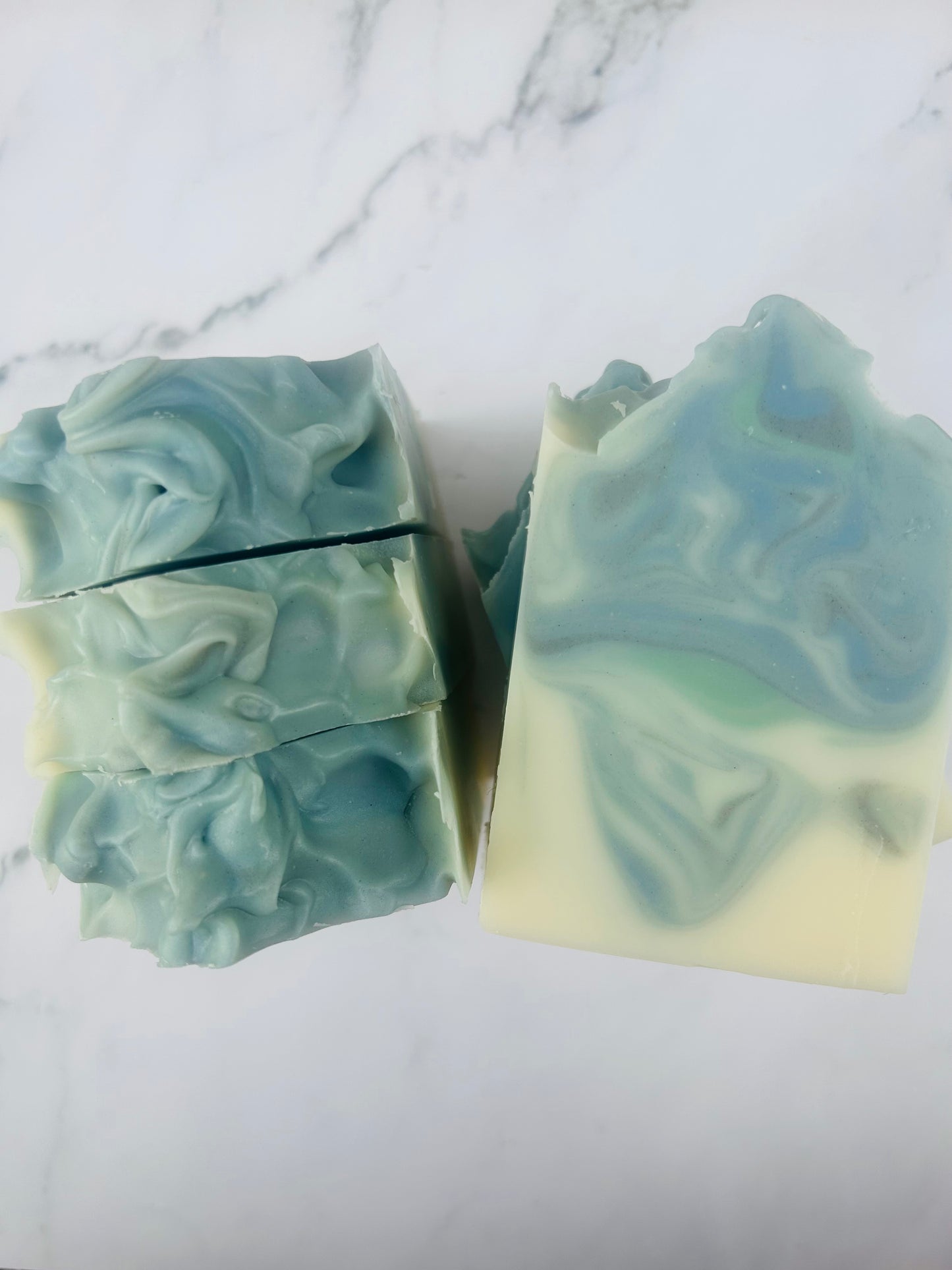 Image of blue, green, and white swirled soap bars with 3 bars stacked on top of one another and 3 bars standing vertically photographed on a white marble background.