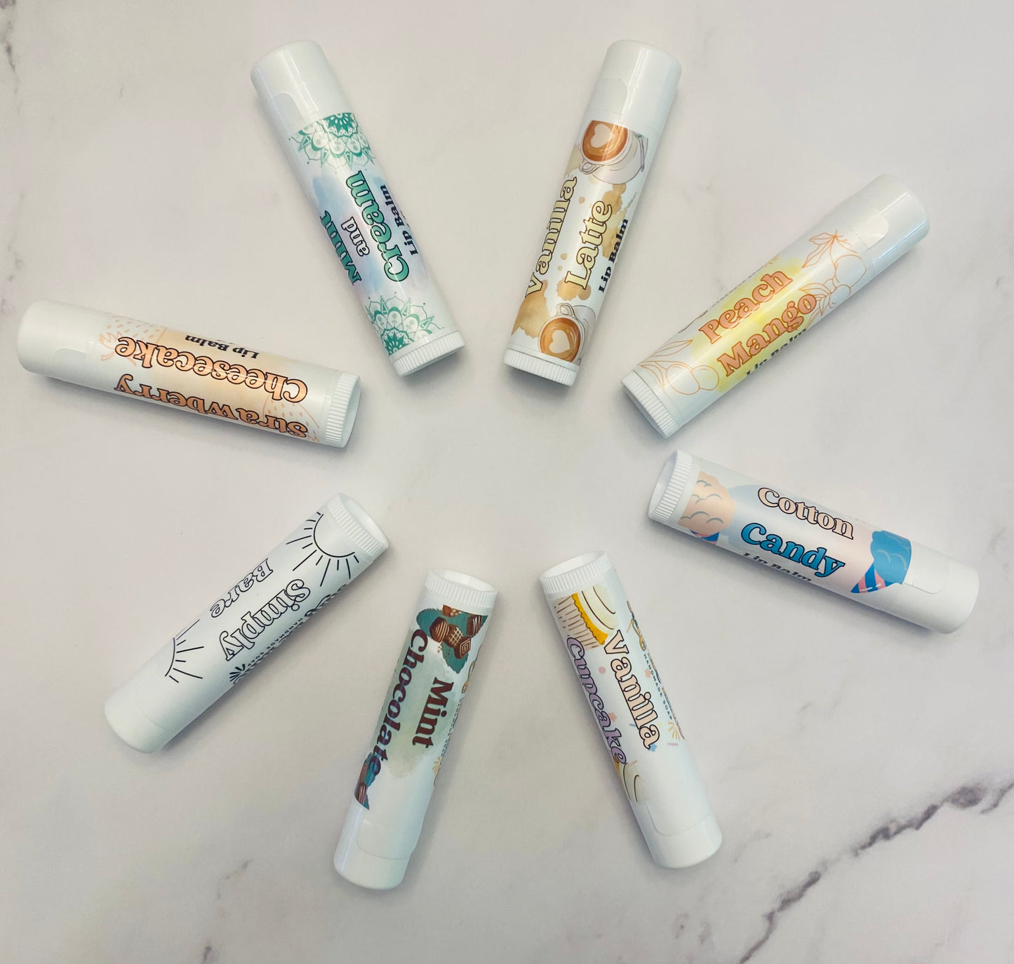 Photo of 8 white lip balm tubes in 8 flavor varieties.: strawberry cheesecake, mint and cream, vanilla latte, peach mango, cotton candy, vanilla cupcake, mint chocolate, and simply bare. Lip balms are displayed in a circle against a white marble background.