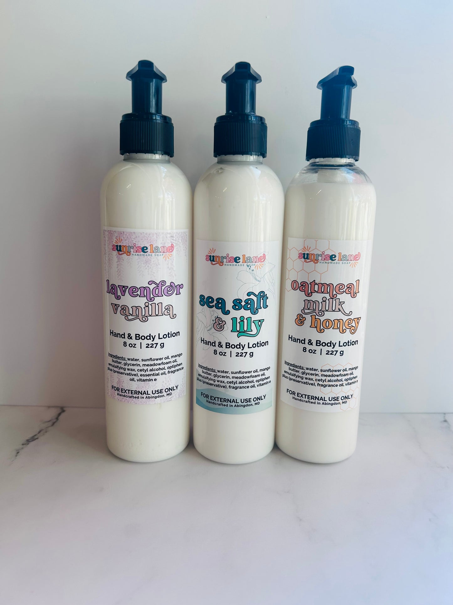 Image of 3 large pump lotion bottles in three scent options: lavender vanilla, sea salt lily, and oatmeal milk honey photographed against a white marble background.