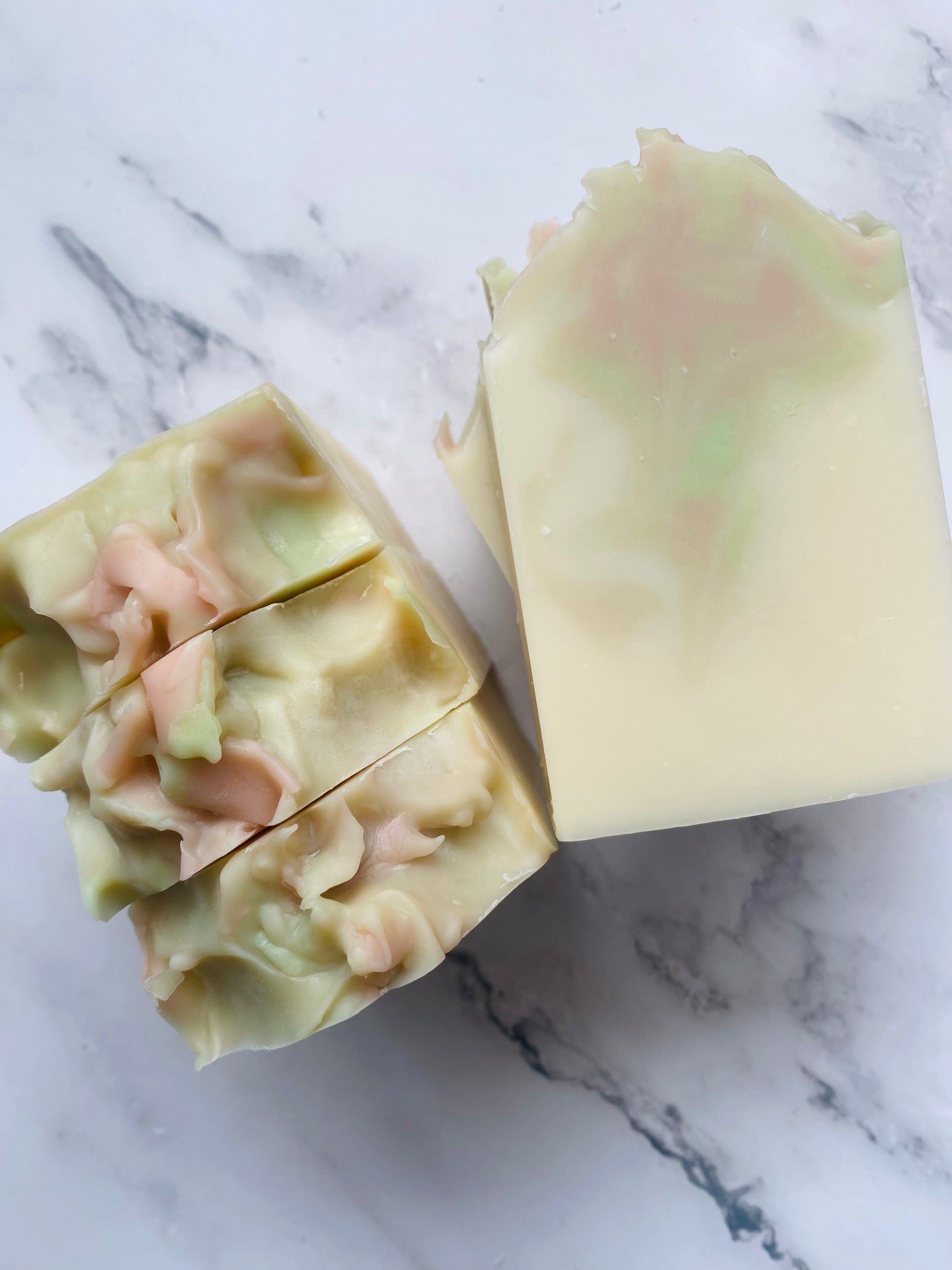 Image of white, pink, and green swirled soap bars with 3 bars stacked on top of one another and 3 bars standing vertically photographed on a white marble background.