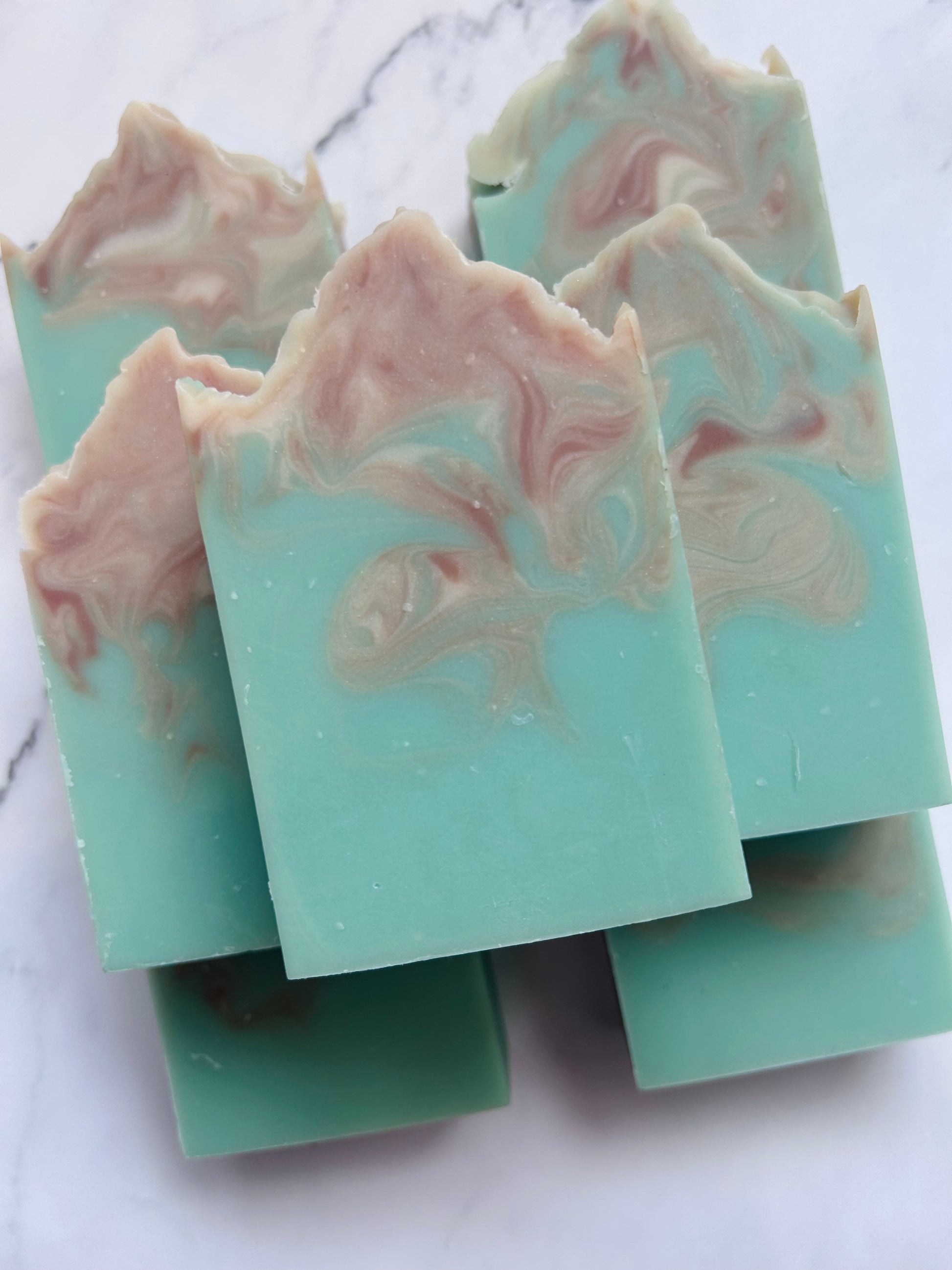 Image of 7 teal, brown, and white swirled soap bars stacked on top of one another photographed on a white marble background.
