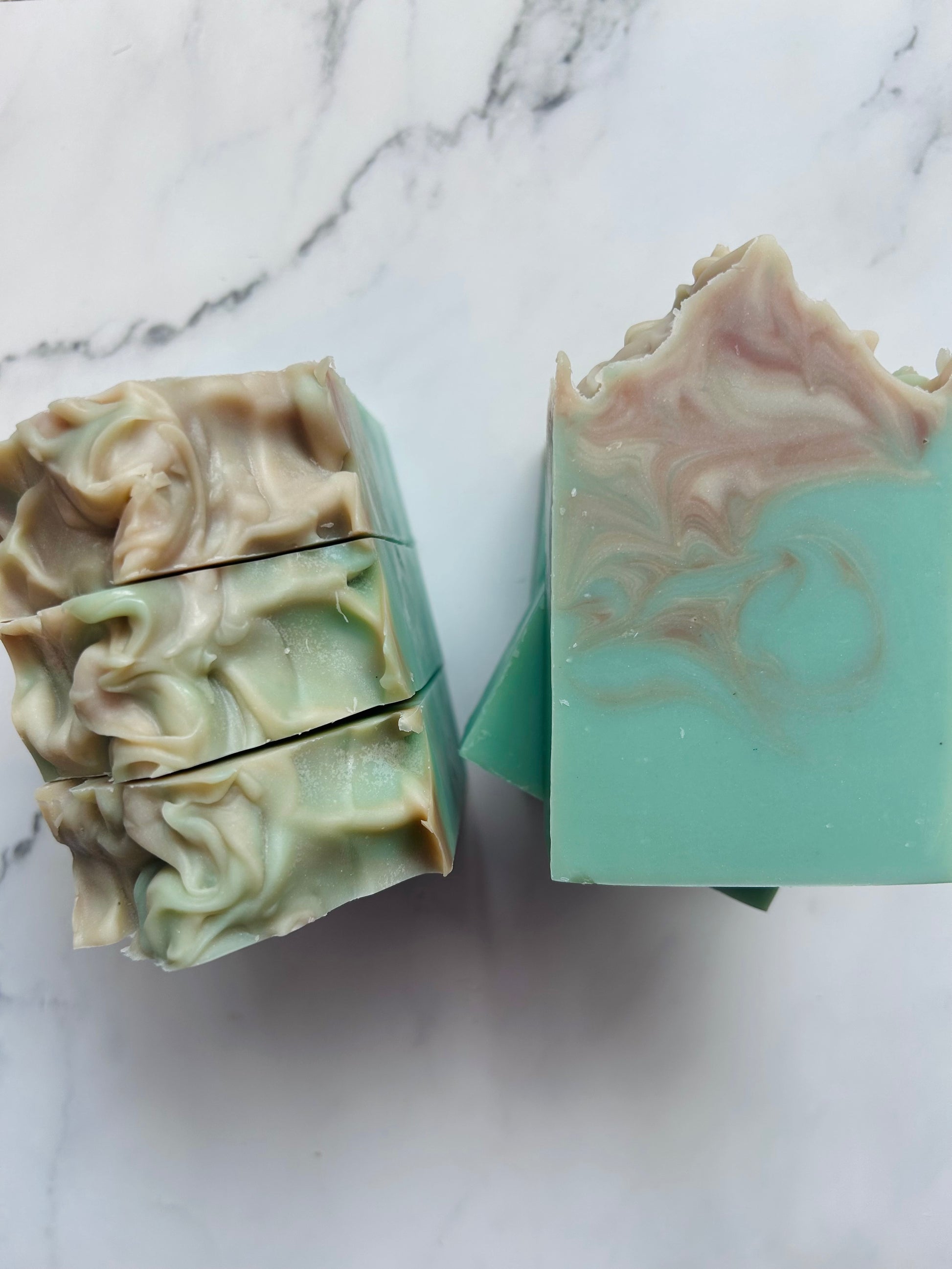 Image of teal, brown, and white swirled soap bars with 3 bars stacked on top of one another and 3 bars standing vertically photographed on a white marble background.