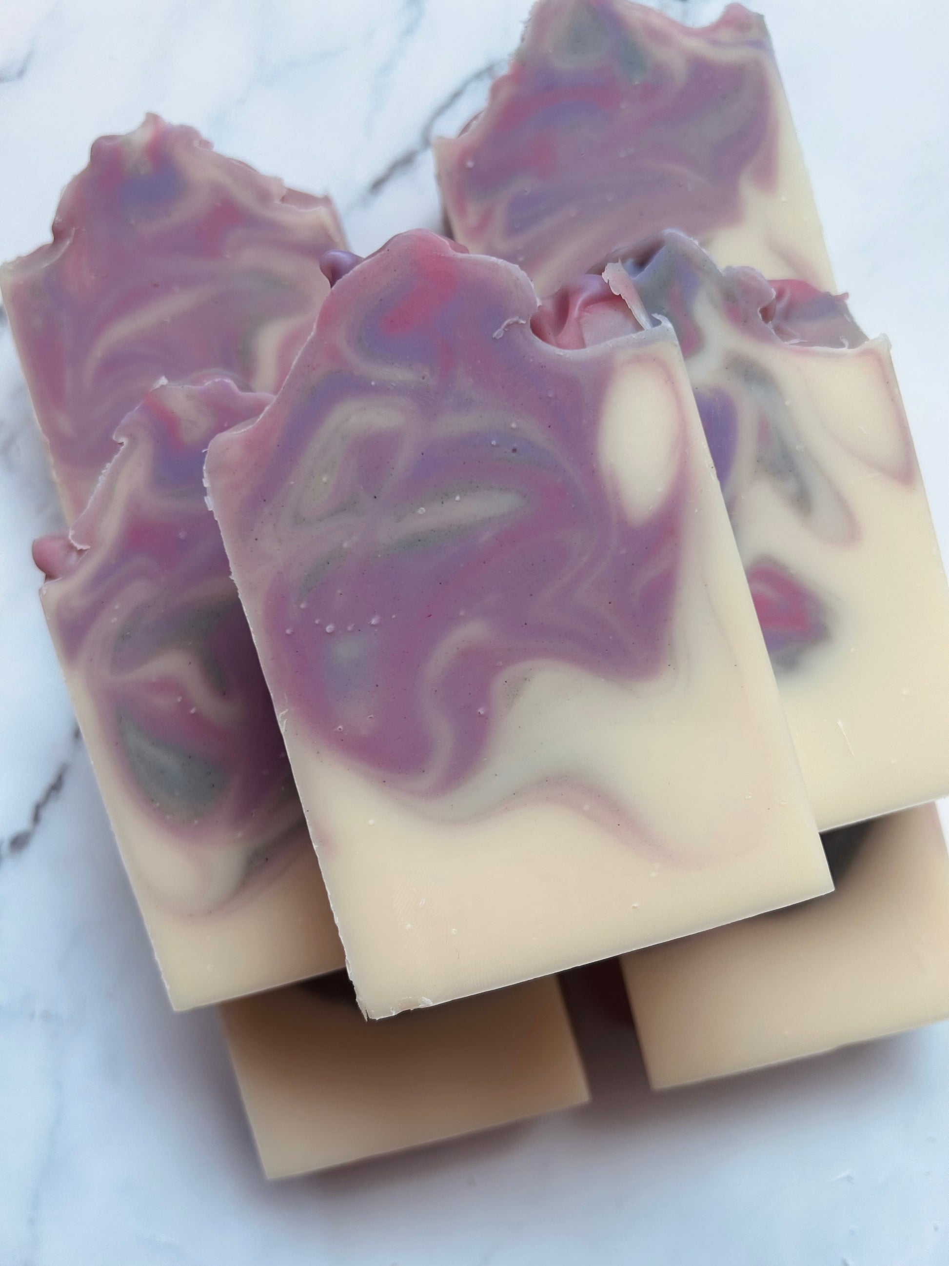 Image of 7 purple, pink, and gray swirled soap bars stacked on top of one another photographed on a white marble background.