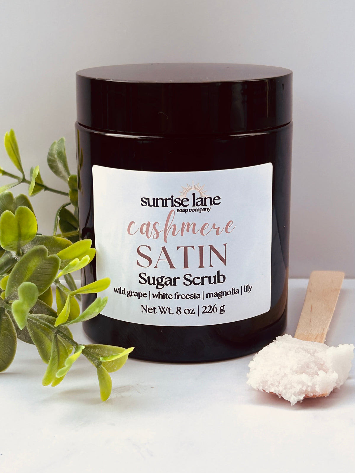 Image of a single black jar containing Cashmere Satin scented sugar scrub. Photographed against a white and gray background with decorative greenery and a sample wooden spoon.