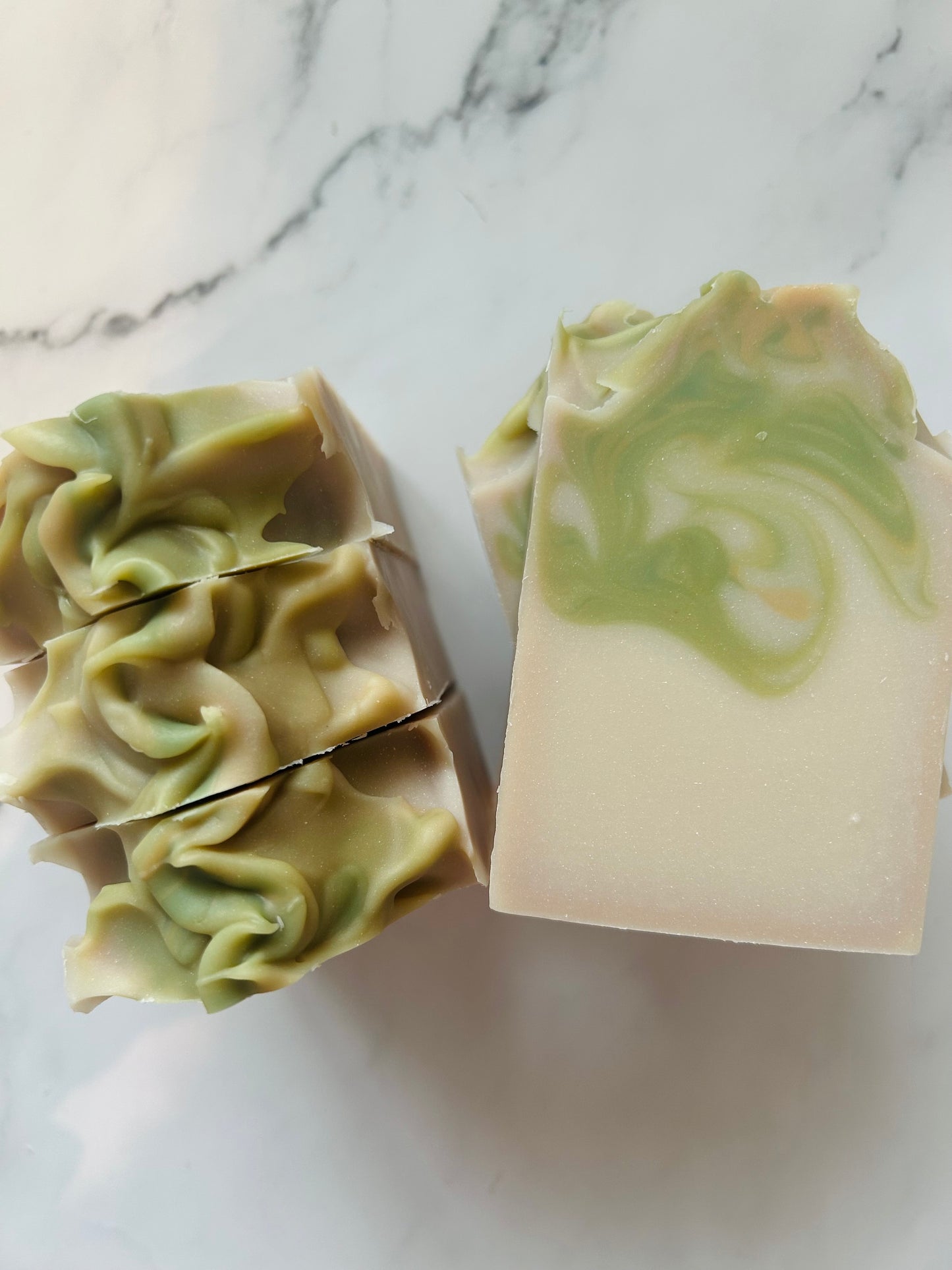 Image of white, green, and gold swirled soap bars with 3 bars stacked on top of one another and 3 bars standing vertically photographed on a white marble background.