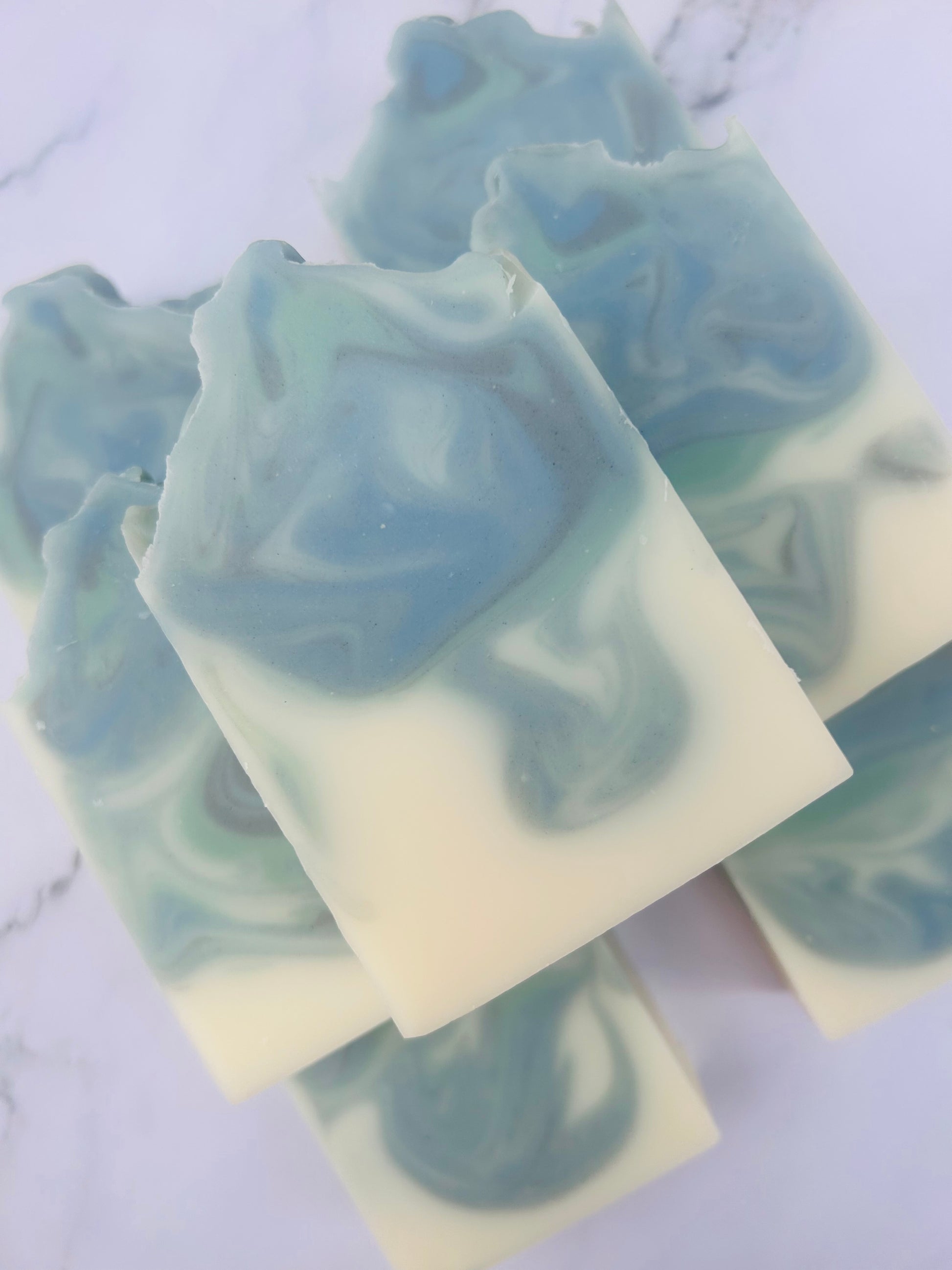 Image of 7 blue, green, and white swirled soap bars stacked on top of one another photographed on a white marble background.