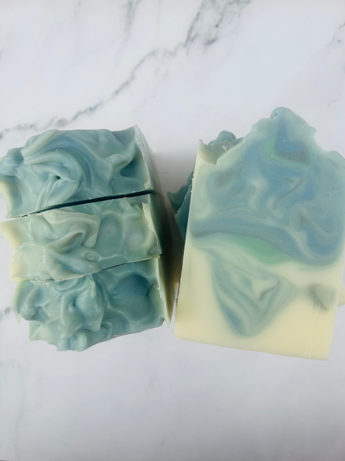 Image of blue, green, and white swirled soap bars with 3 bars stacked on top of one another and 3 bars standing vertically photographed on a white marble background.