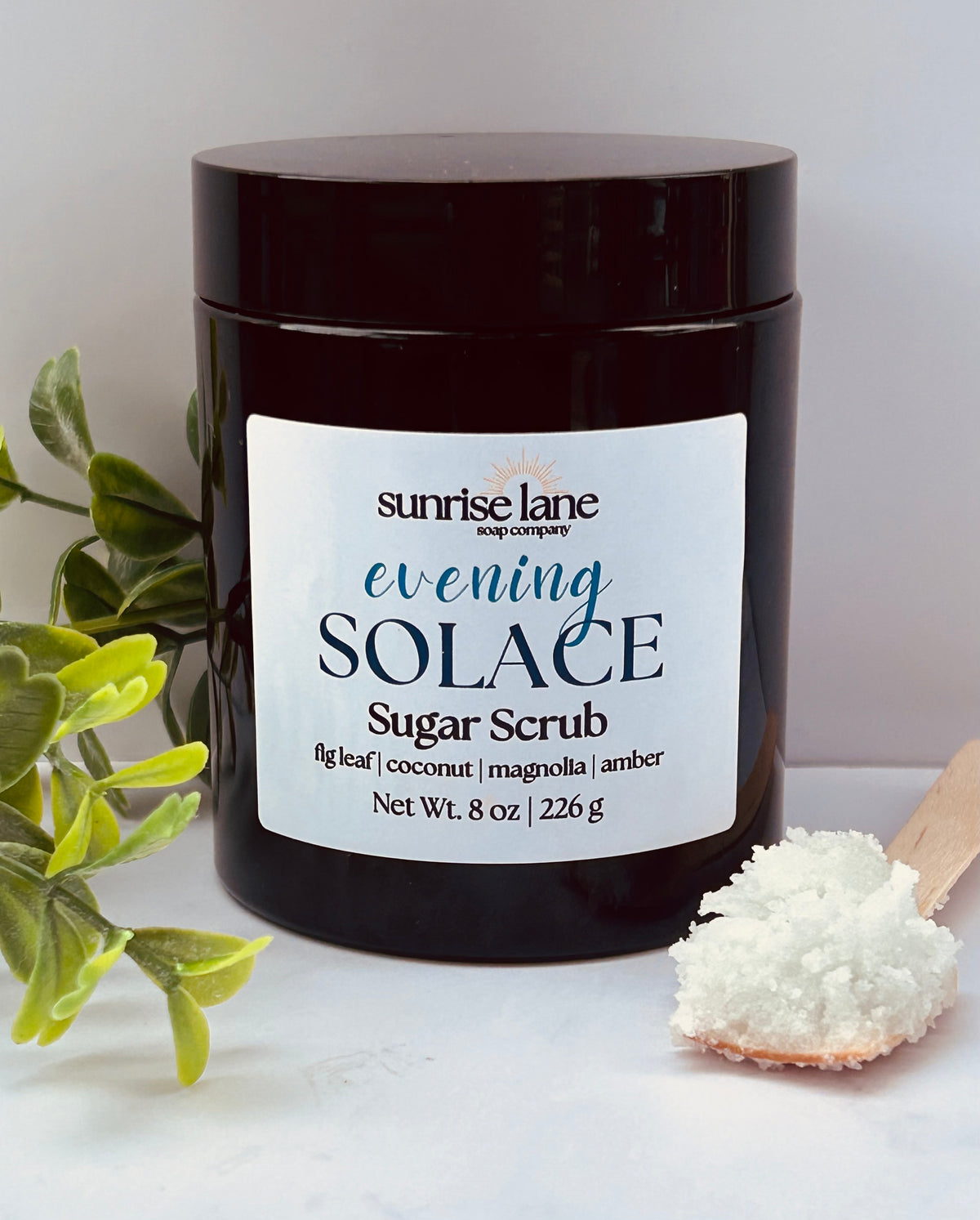 Image of a single black jar containing Evening Solace scented sugar scrub. Photographed against a white and gray background with decorative greenery and a sample wooden spoon.