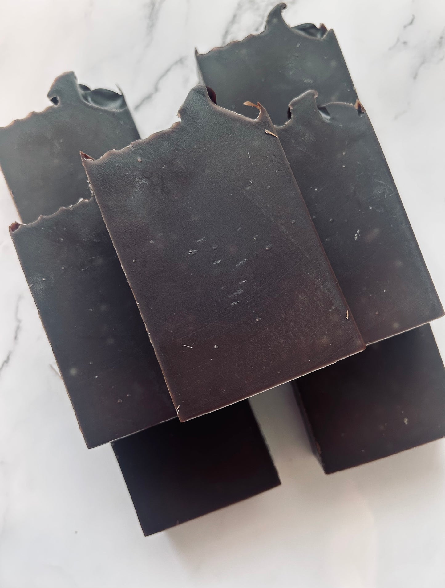 Image of 7 dark brown soap bars stacked on top of one another photographed on a white marble background.