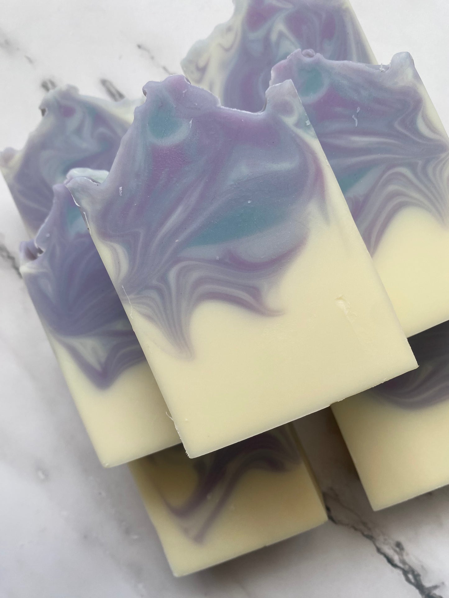 Image of 7 white, purple, and blue swirled soap bars stacked on top of one another photographed on a white marble background.