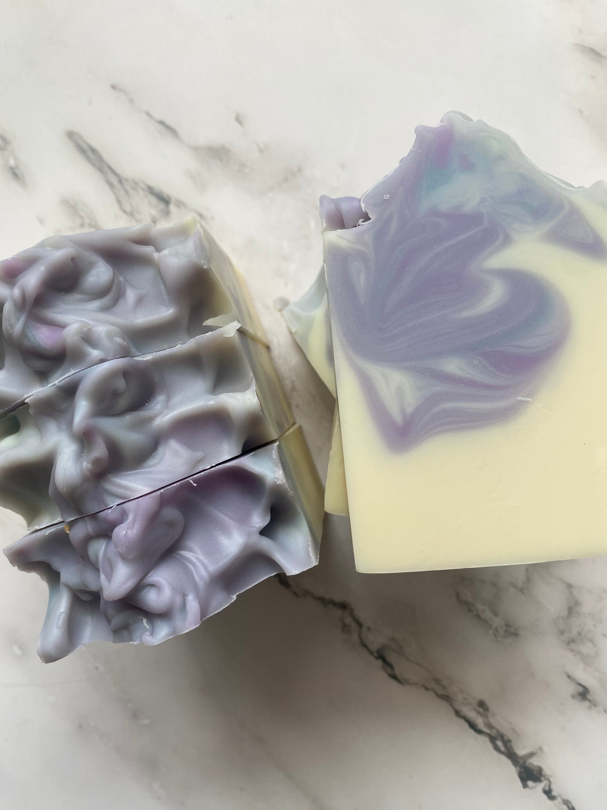 Image of white, purple, and blue swirled soap bars with 3 bars stacked on top of one another and 3 bars standing vertically photographed on a white marble background.