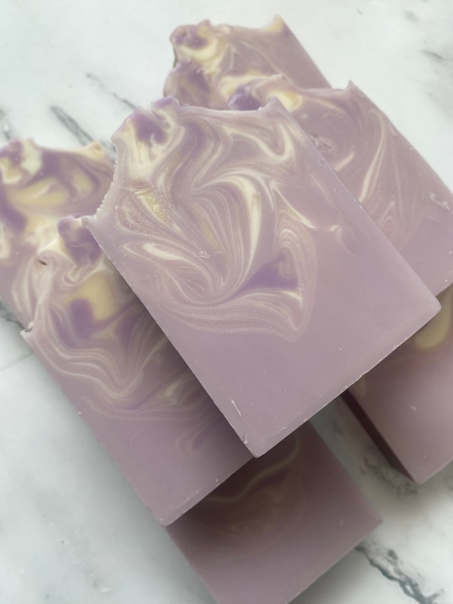 Image of 7 purple, white, and tan swirled soap bars stacked on top of one another photographed on a white marble background.
