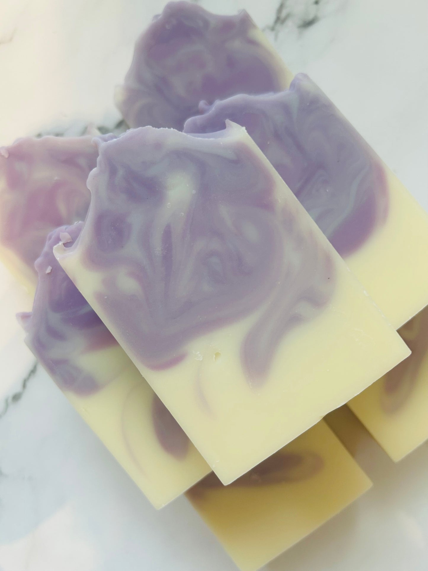 Image of 7 white and purple swirled soap bars stacked on top of one another photographed on a white marble background.