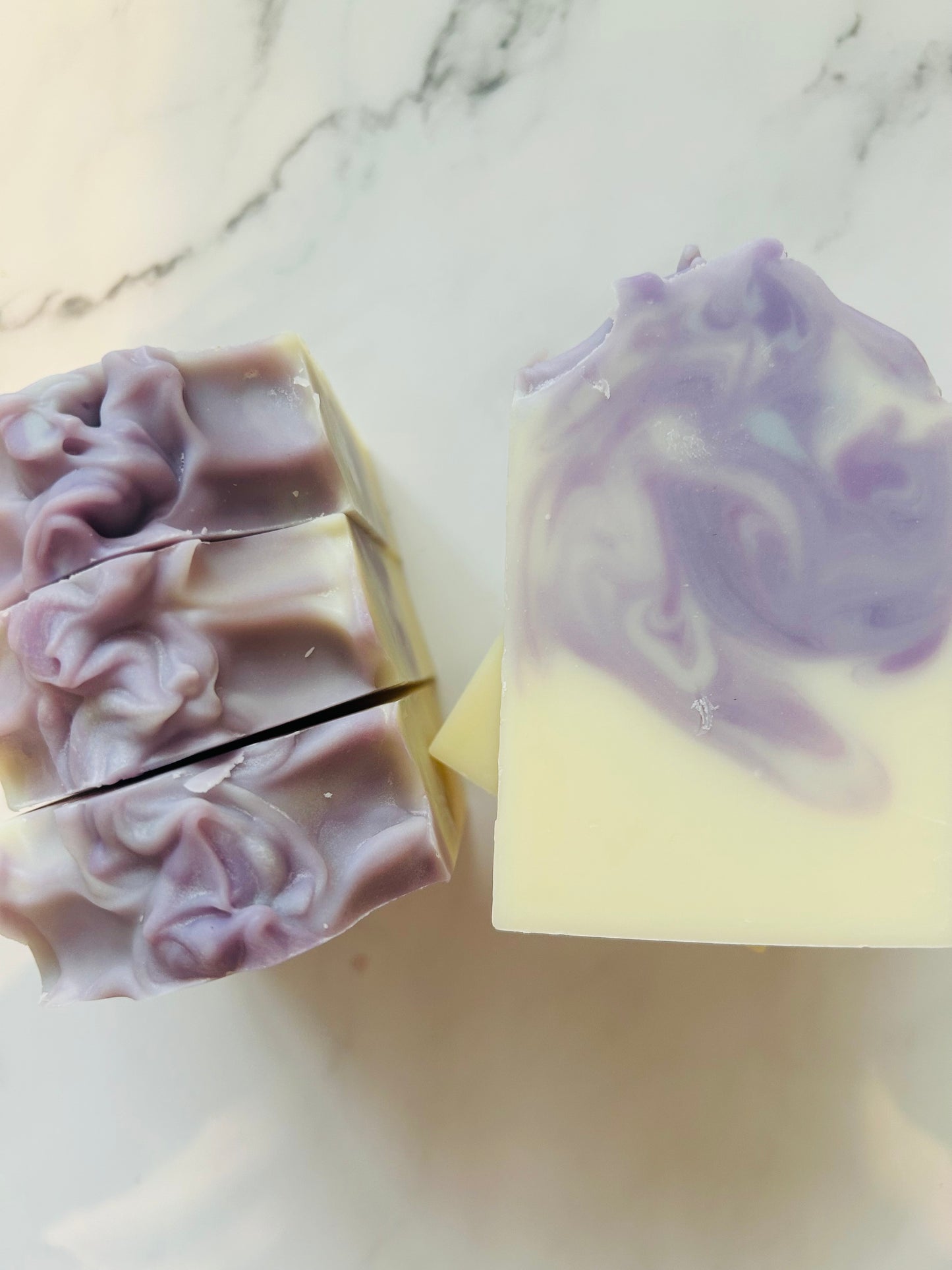 Image of white and purple swirled soap bars with 3 bars stacked on top of one another and 3 bars standing vertically photographed on a white marble background.