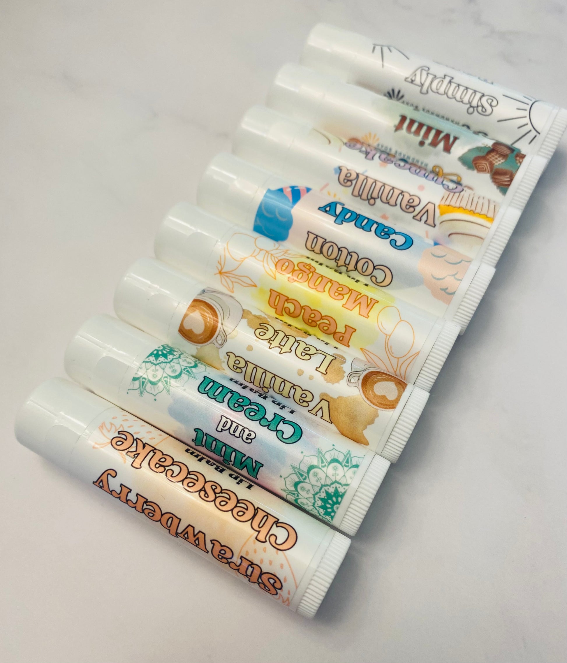 Photo of 8 white lip balm tubes in 8 flavor varieties.: strawberry cheesecake, mint and cream, vanilla latte, peach mango, cotton candy, vanilla cupcake, mint chocolate, and simply bare. Lip balms are displayed diagonally in a row against a white background.