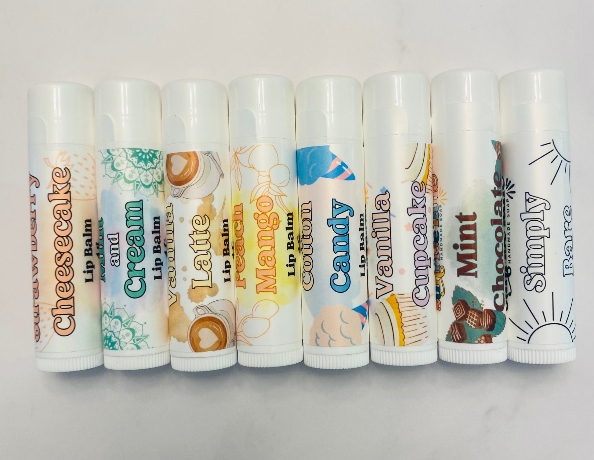 Photo of 8 white lip balm tubes in 8 flavor varieties.: strawberry cheesecake, mint and cream, vanilla latte, peach mango, cotton candy, vanilla cupcake, mint chocolate, and simply bare. Lip balms are displayed horizontally in a row against a white background.