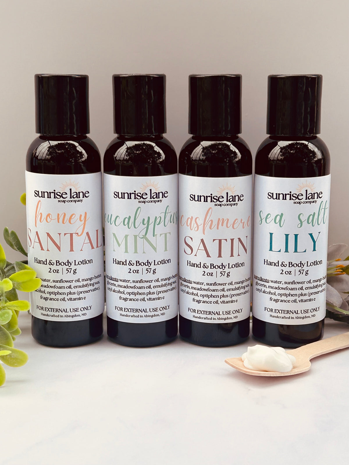 Image of four small, black lotion bottles containing four scent varieties. Scents include: Honey Santal, Eucalyptus Mint, Cashmere Satin, and Sea Salt Lily. Photographed against a white and gray background with decorative greenery and a sample wooden spoon.