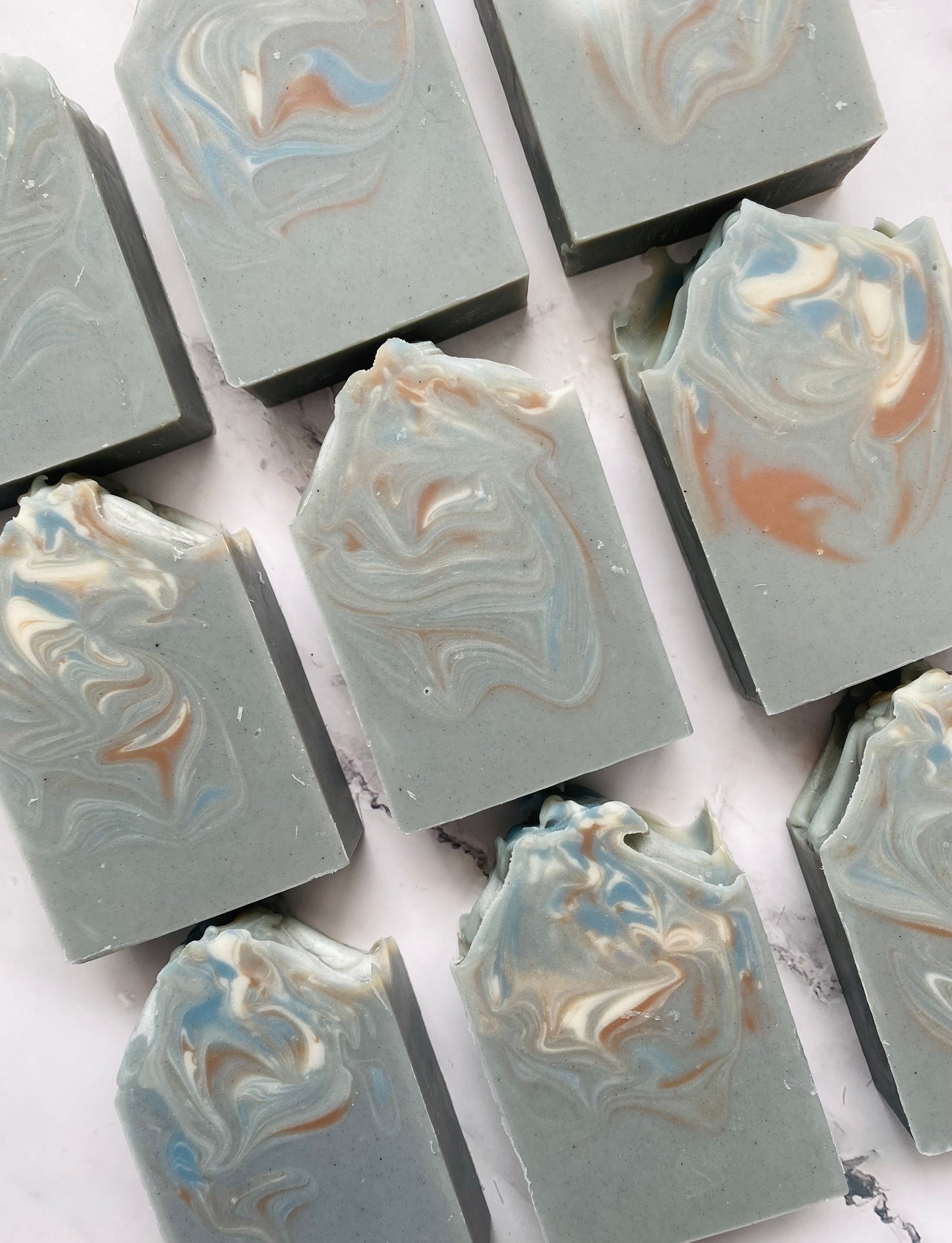 Image of 9 gray, blue, white, and brown swirled soap bars photographed vertically on a white marble background.