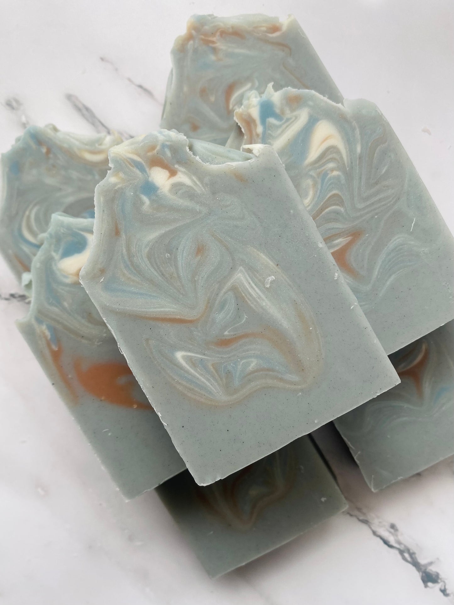 Image of 7 gray, blue, white, and brown swirled soap bars stacked on top of one another photographed on a white marble background.
