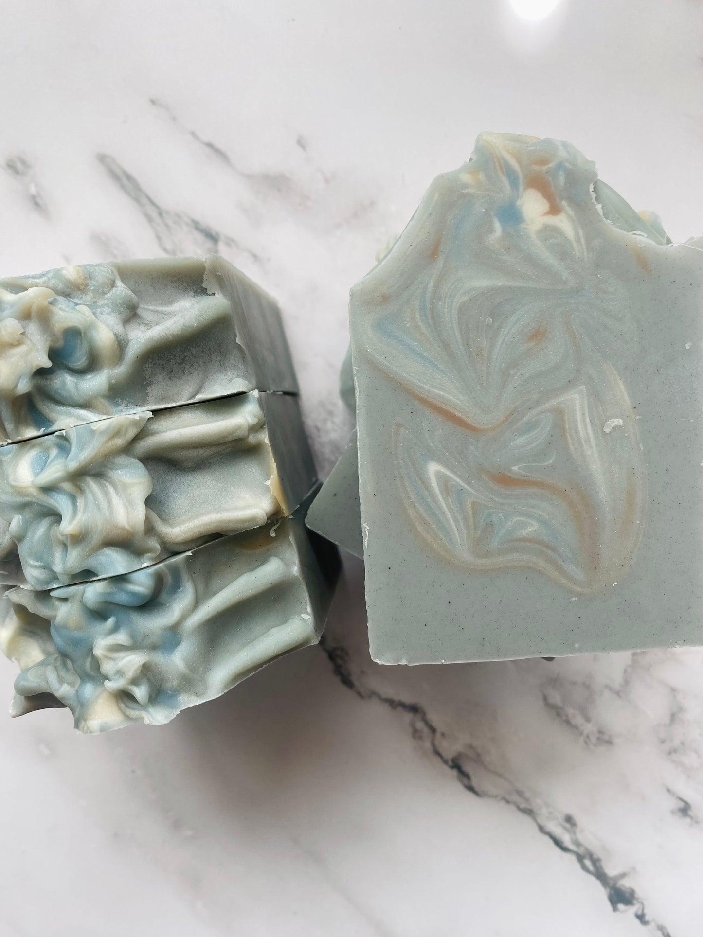 Image of gray, blue, white, and brown swirled soap bars with 3 bars stacked on top of one another and 3 bars standing vertically photographed on a white marble background.