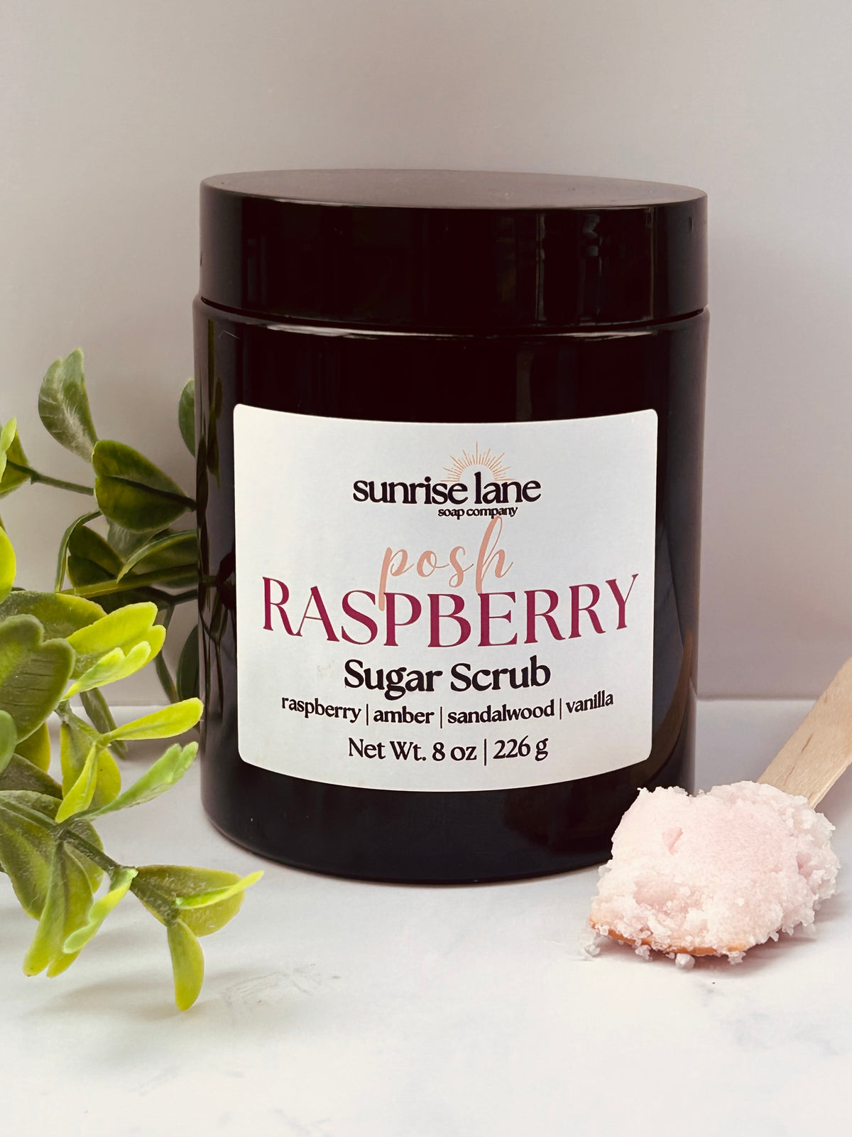 Image of a single black jar containing Posh Raspberry scented sugar scrub. Photographed against a white and gray background with decorative greenery and a sample wooden spoon.