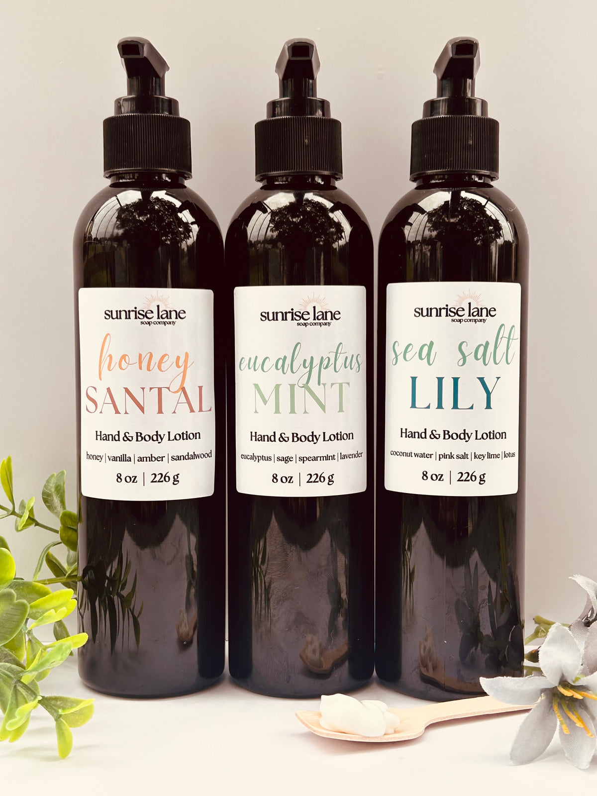 Image of three large black pump lotion bottles containing three scent varieties. Scents include: Honey Santal, Eucalyptus Mint, and Sea Salt Lily. Photographed against a white and gray background with decorative greenery and a sample wooden spoon.
