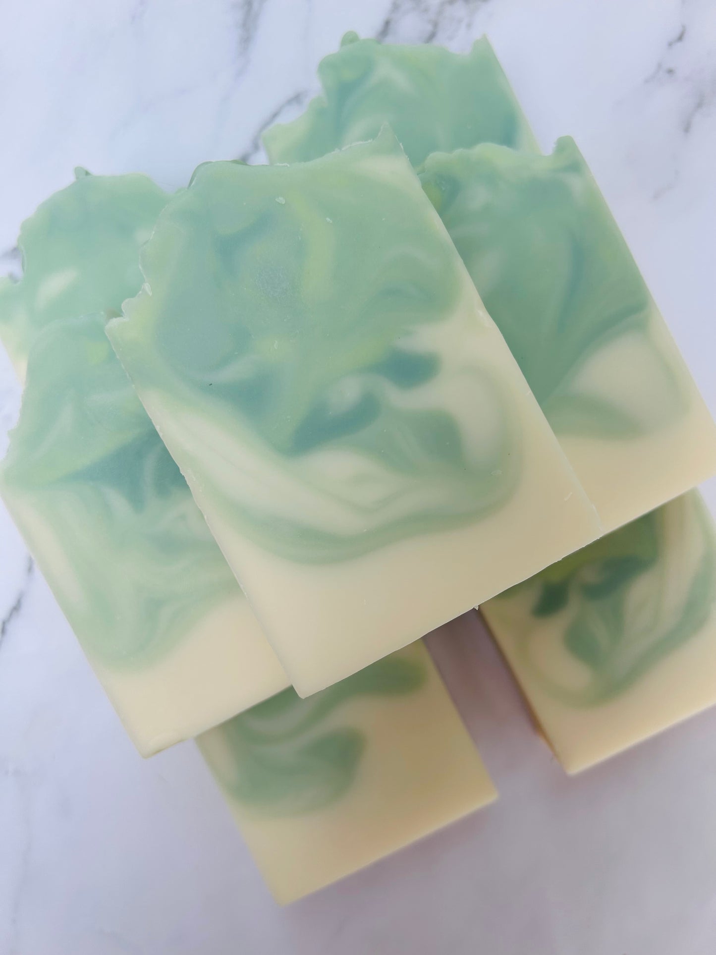 Image of 7 green and white swirled soap bars stacked on top of one another photographed on a white marble background.