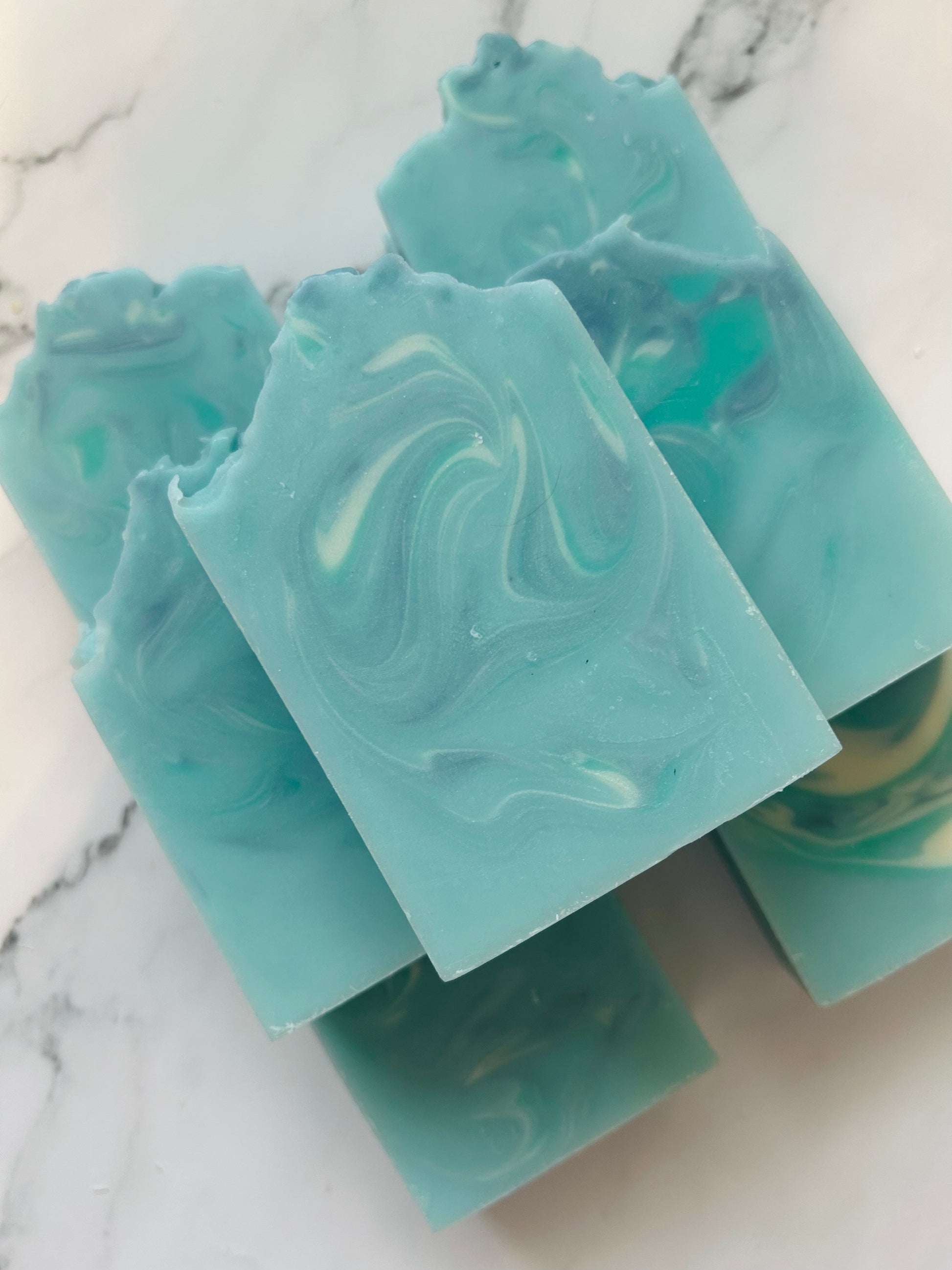 Image of 7 blue, white, and teal swirled soap bars stacked on top of one another photographed on a white marble background.