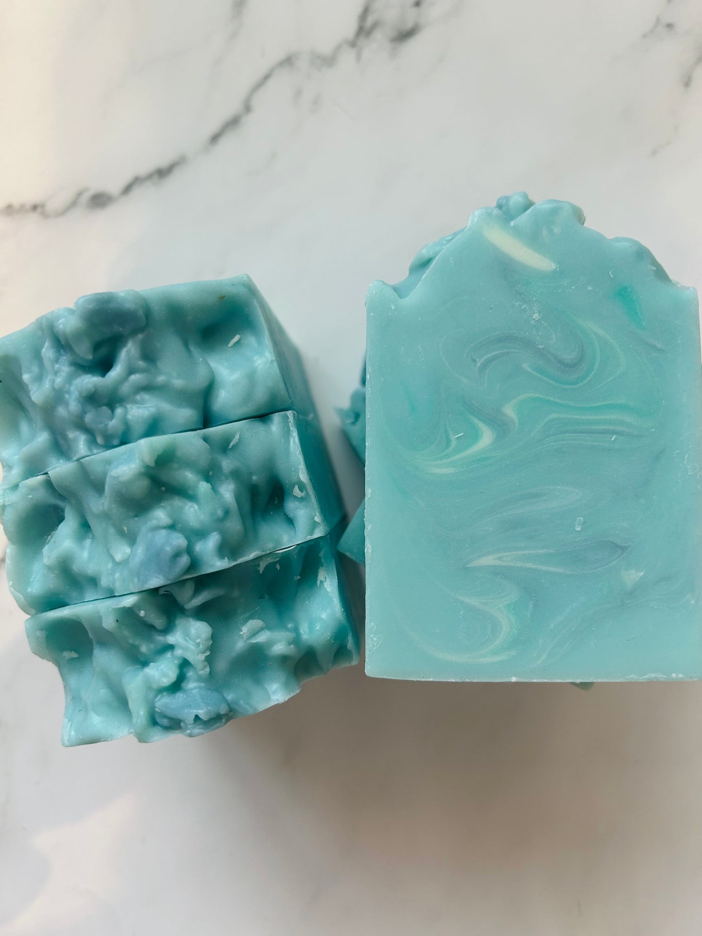 Image of blue, white, and teal swirled soap bars with 3 bars stacked on top of one another and 3 bars standing vertically photographed on a white marble background.