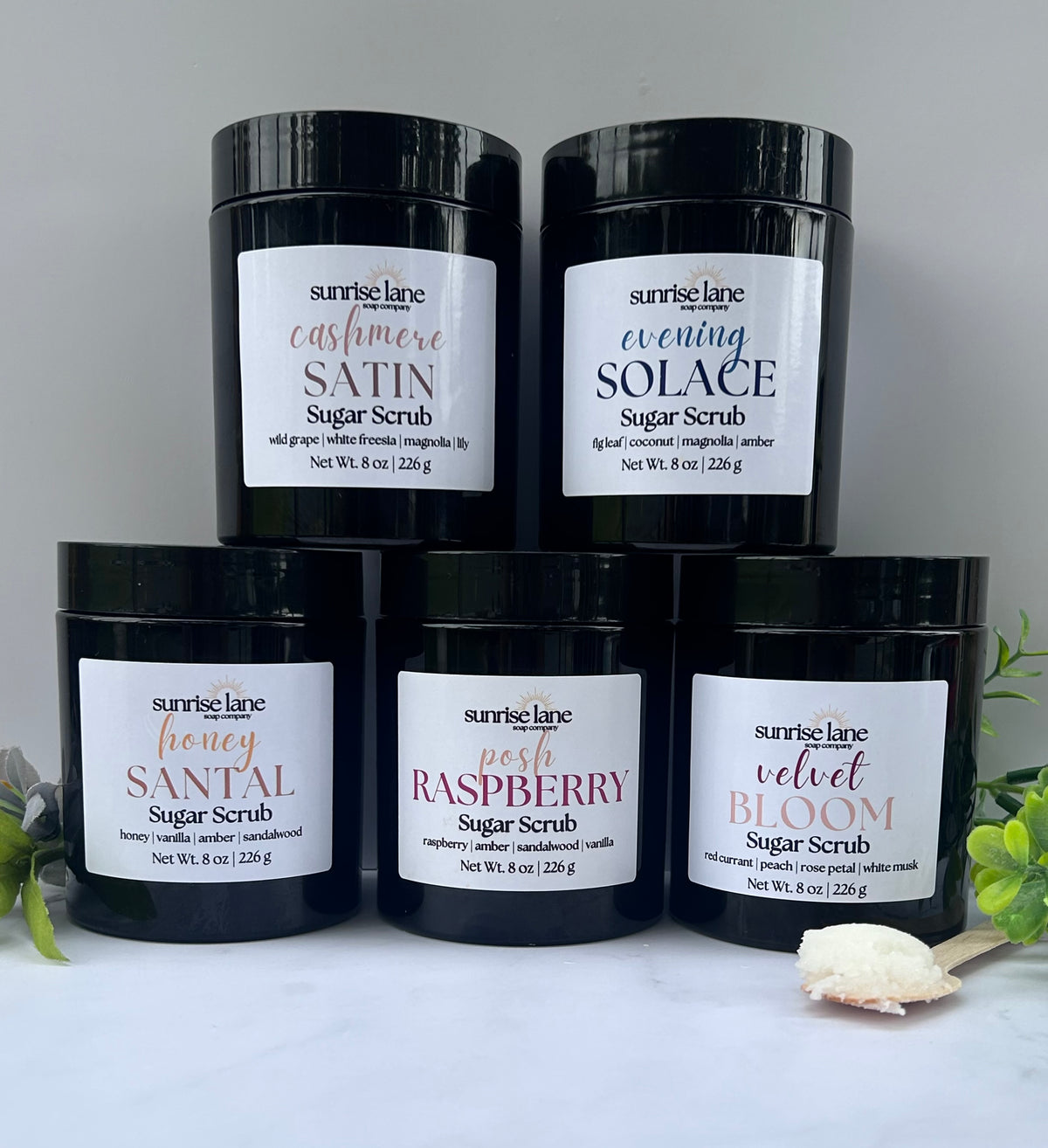 Image of 5 stacked black jars containing 5 varieties of sugar scrub. Scents include: Cashmere Satin, Evening Solace, Honey Santal, Posh Raspberry, and Velvet Bloom. Jars are stacked two on top of three against a white and gray background with decorative greenery and a sample wooden spoon.