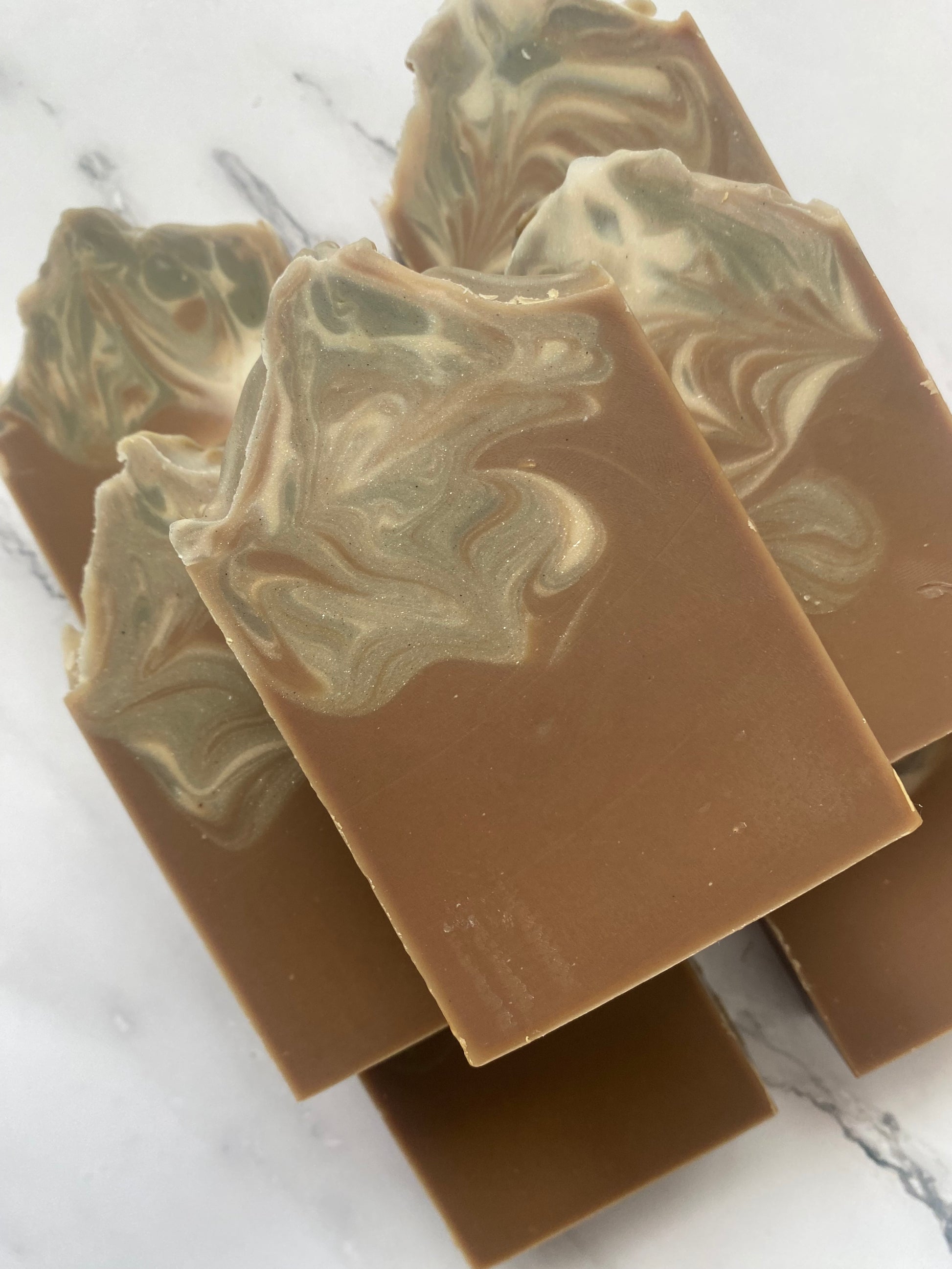Image of 7 brown, white, and gray swirled soap bars stacked on top of one another photographed on a white marble background.